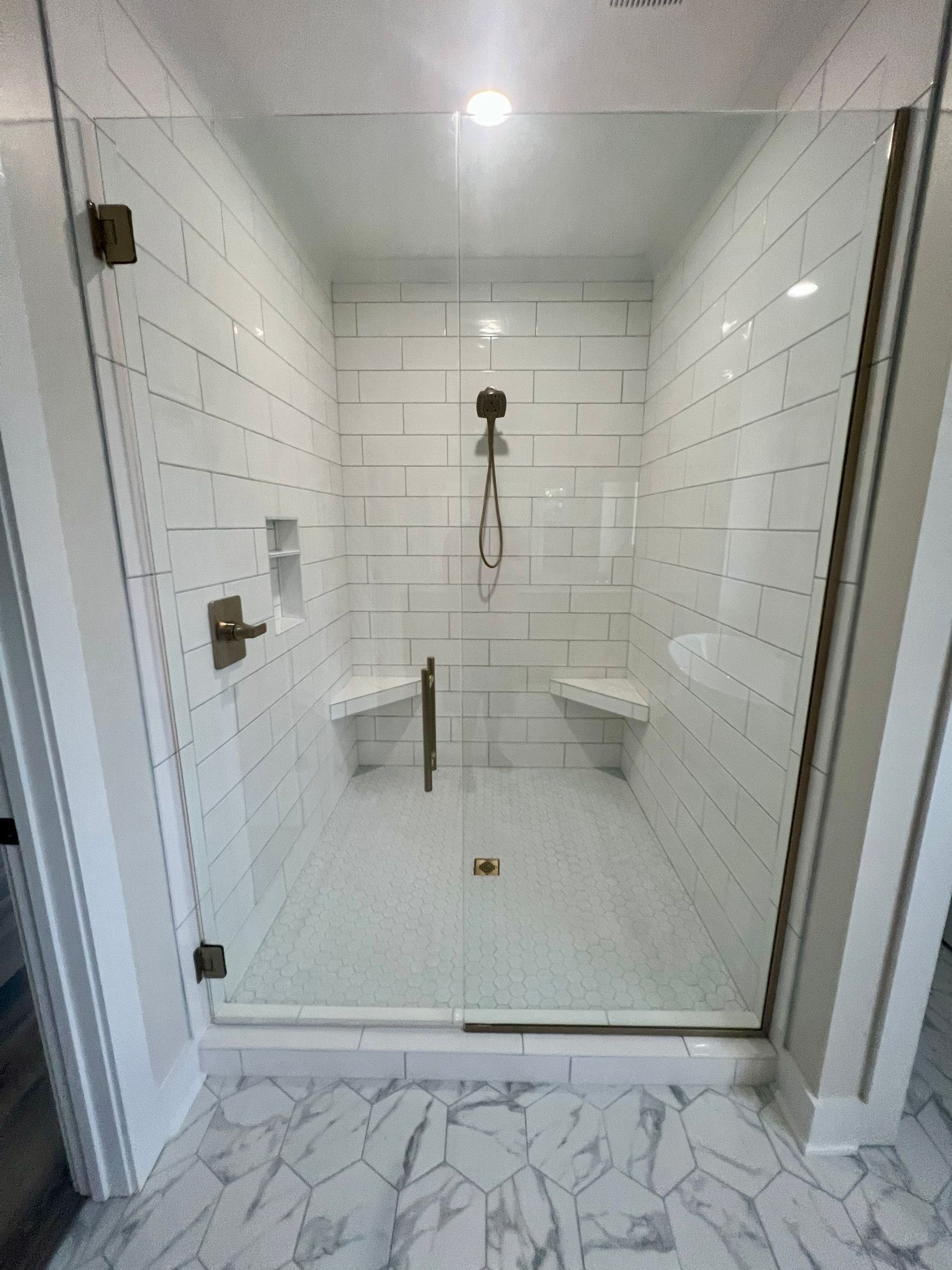 Shower with glass doors and white tiled walls. Features built-in shelves and two corner seats.