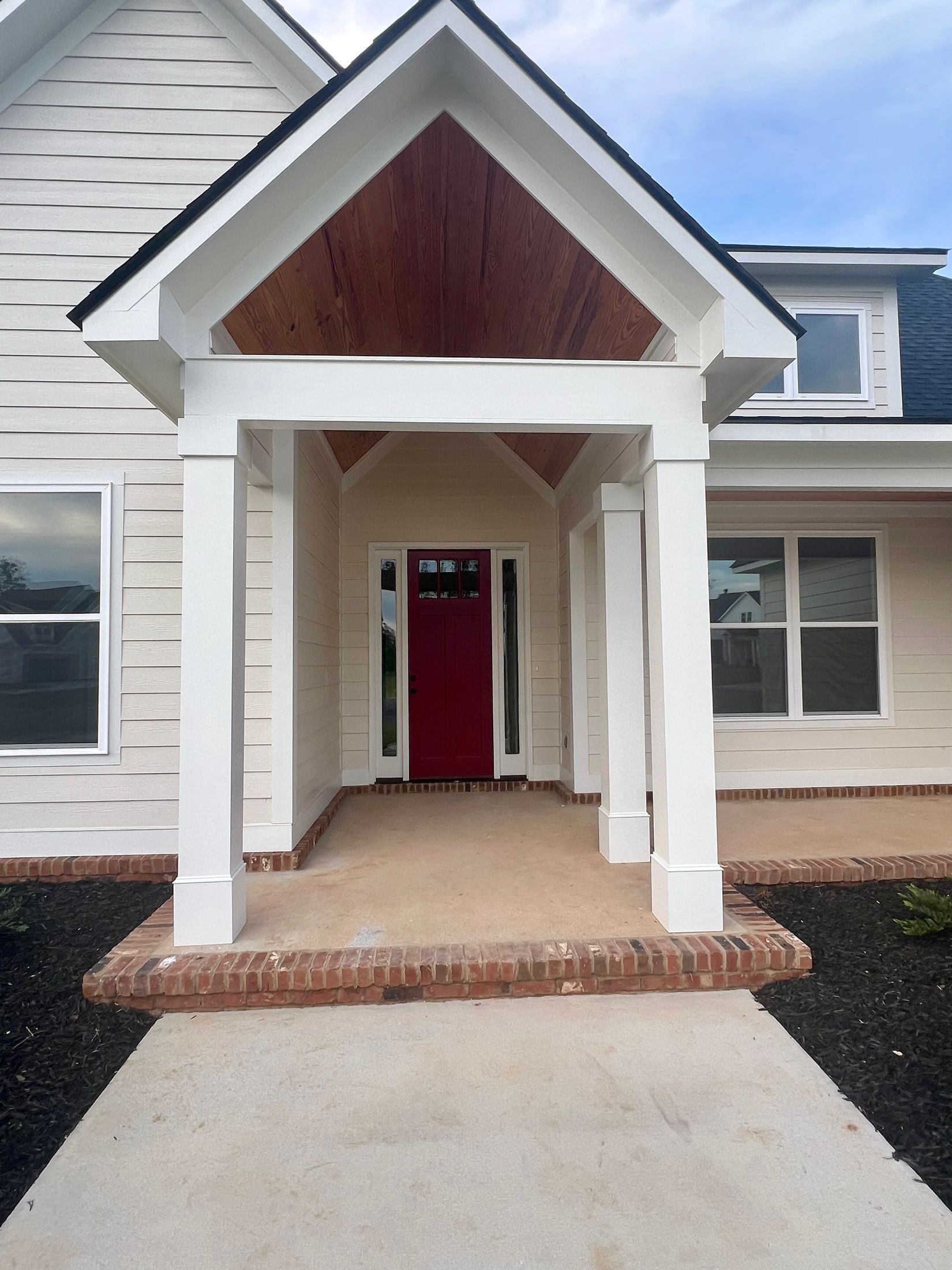 Red front door under a white porch with brick base and concrete walkway.