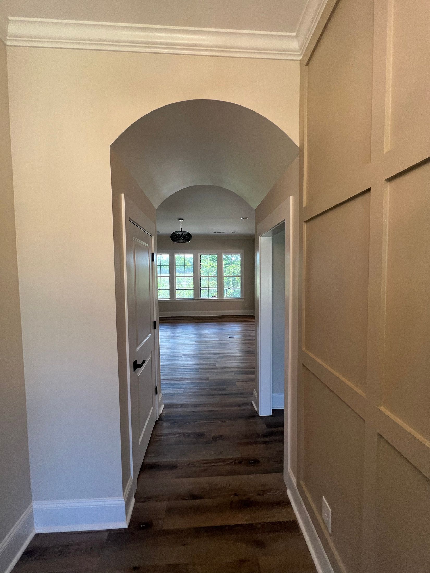 Hallway with arched doorway leading to a room with windows and hardwood floors. Walls are beige and have paneling.