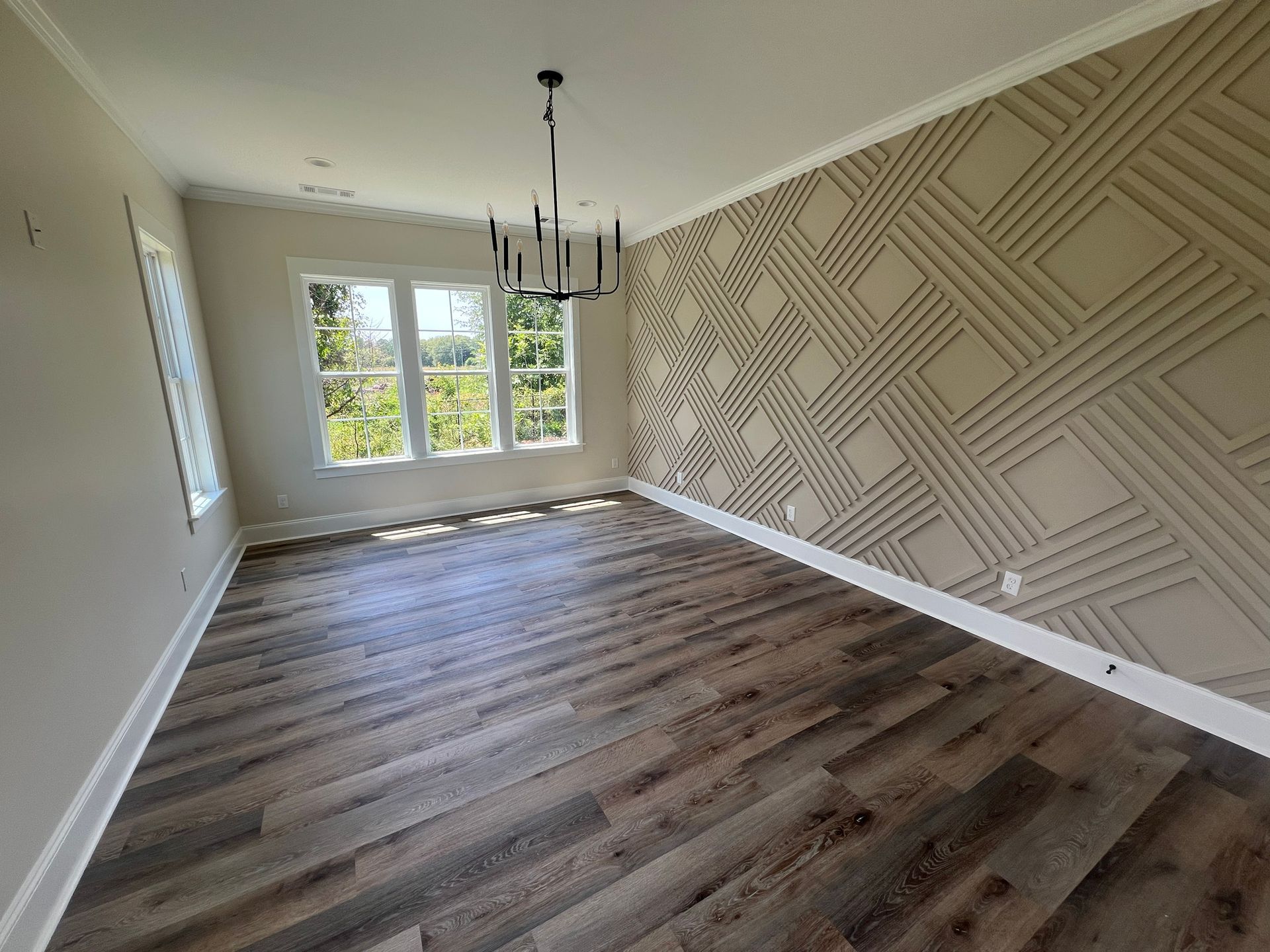 Empty room with wood flooring, patterned accent wall, chandelier, and windows overlooking greenery.