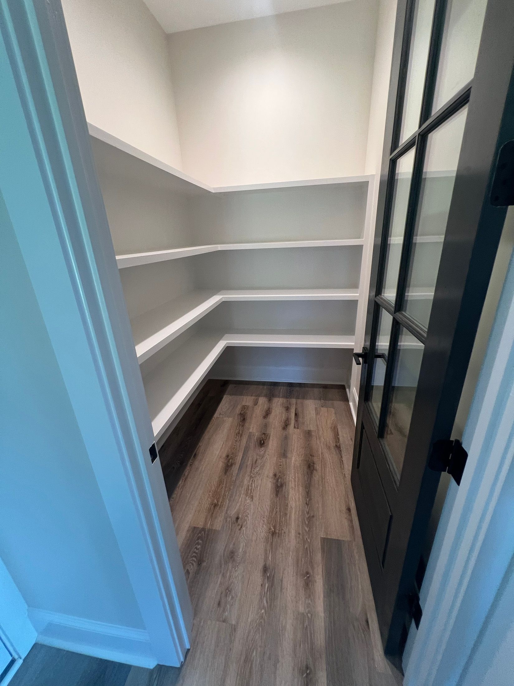 A walk-in pantry with white shelves and a dark-framed glass door. The floor is light wood.