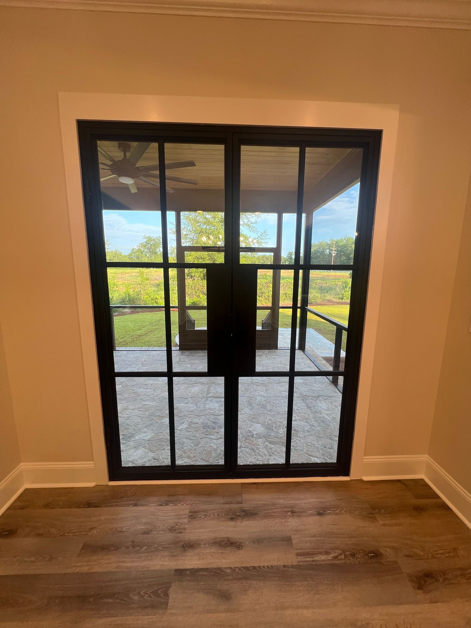 Black-framed glass doors open to a patio. Beige walls and gray floor. Green trees in the distance.