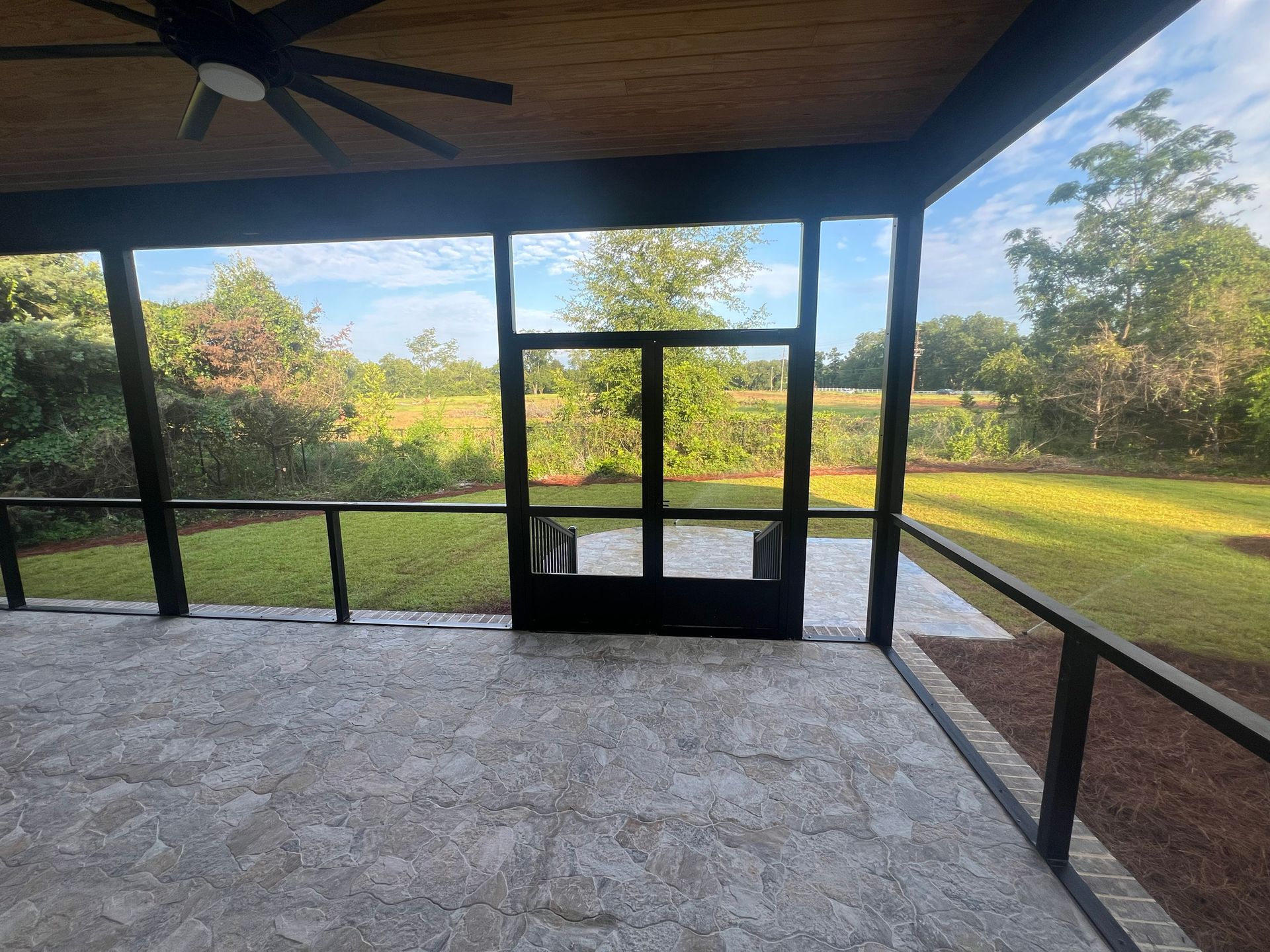 Screened-in porch with view of yard and trees; tile floor, black frame, ceiling fan.
