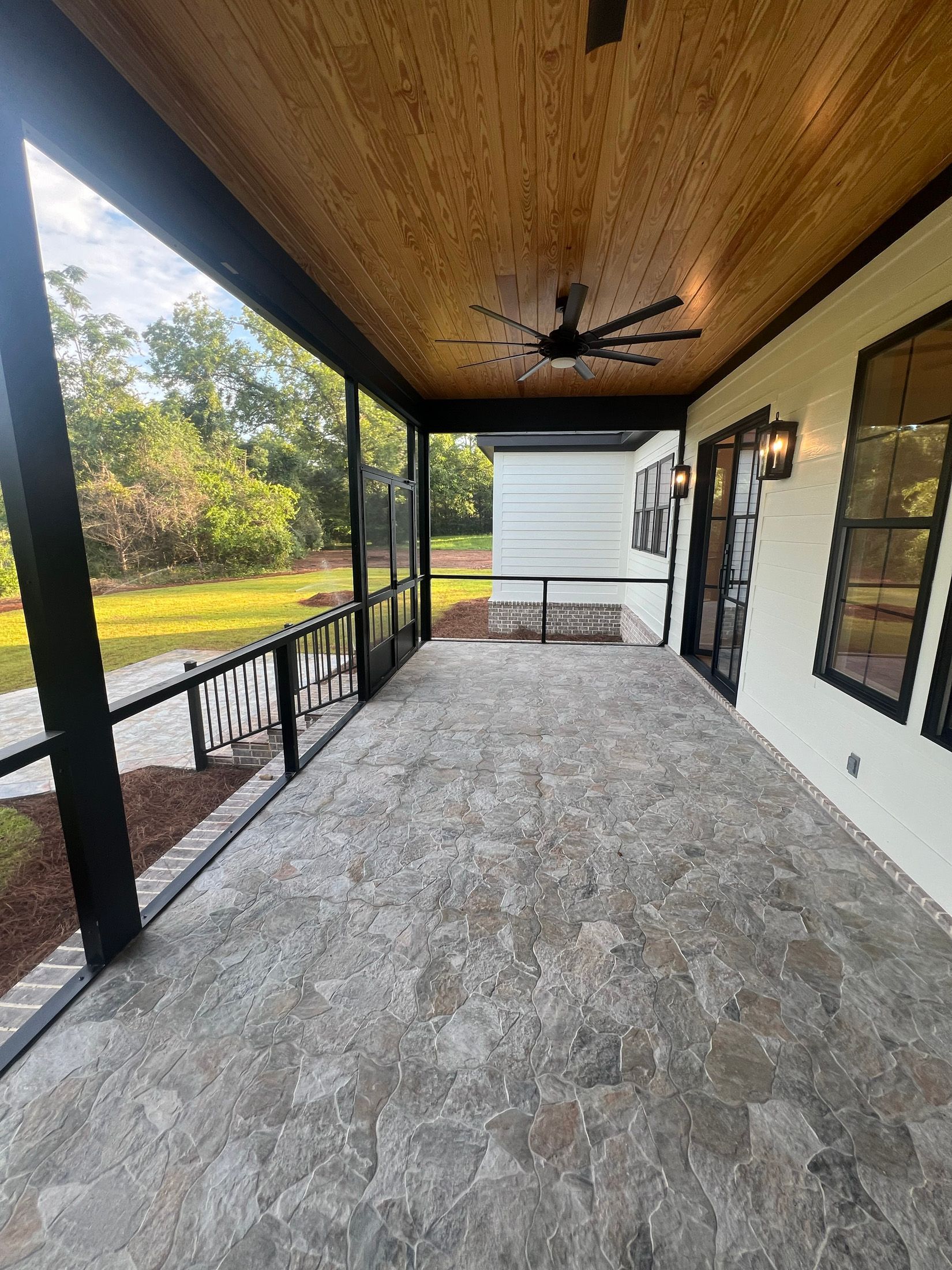 Covered porch with stone floor, black trim, and wood ceiling. The porch overlooks a grassy yard.
