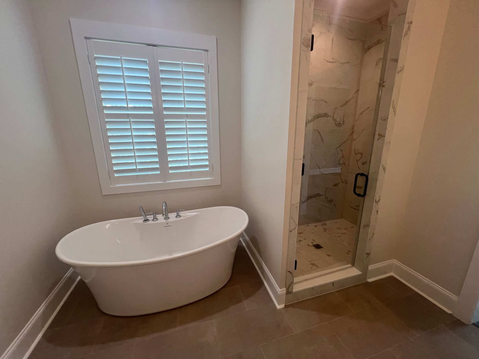 Bathroom with a white oval bathtub, shower, and a window with shutters. The walls are neutral.