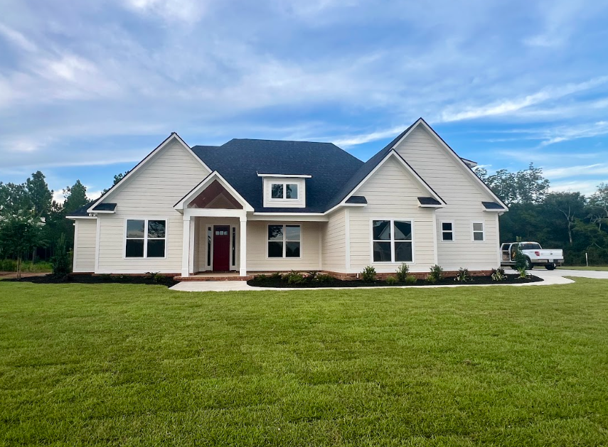 Beige house with black roof, red front door, white trim, and a green lawn under a cloudy sky.