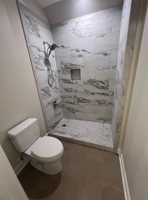 Small bathroom with a toilet and marble-tiled shower, featuring a recessed shelf and rain shower head.