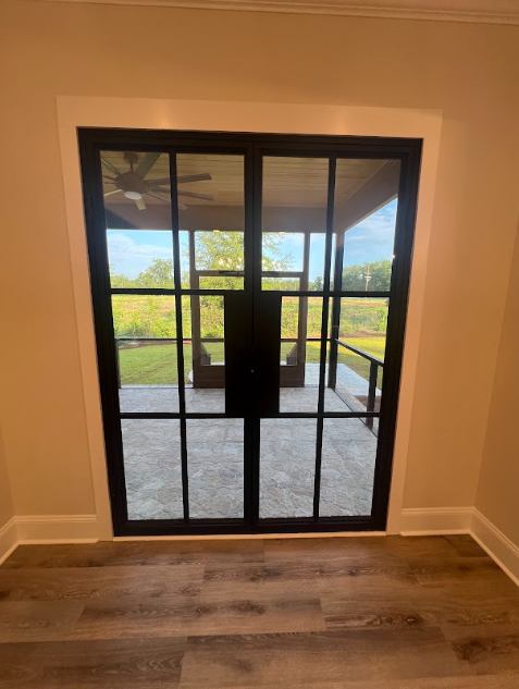 Black framed glass doors, view of outdoor patio and green landscape. Beige walls, light wood floor.