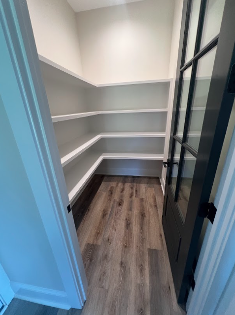 Small pantry with wooden floors, white shelves, and a glass-paneled black door.