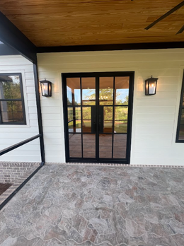 Black-framed glass double doors on a beige house, flanked by two wall-mounted lanterns and windows, with a stone patio.