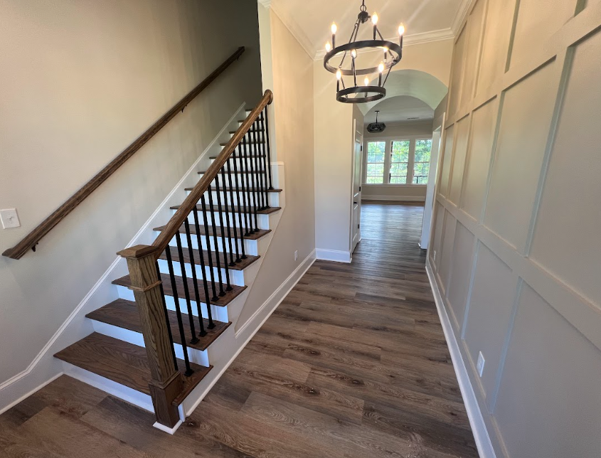 Entryway with staircase, wood floors, and paneling. Black iron spindles, brown banister, and chandelier.