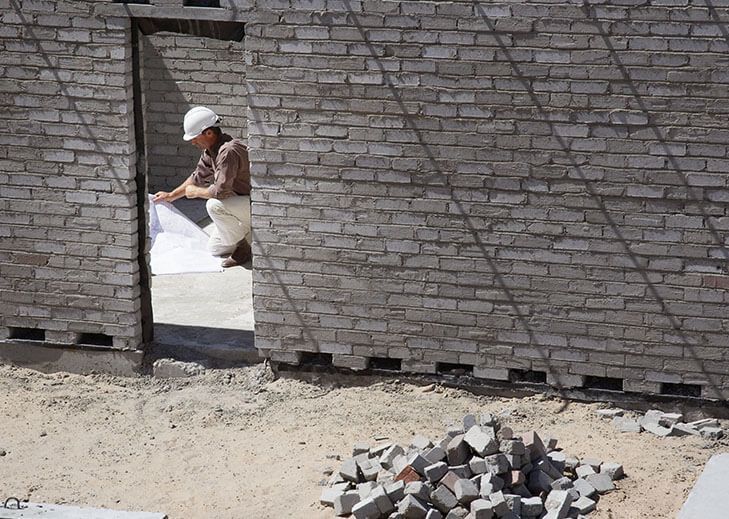Construction worker in a hard hat studying blueprints at an unfinished brick wall doorway.