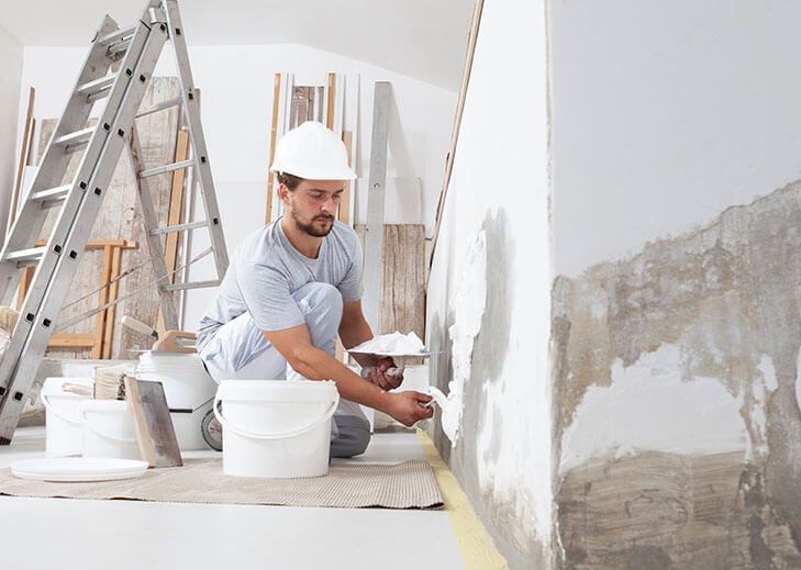 Man in hardhat plastering a wall in a room, with buckets, tools, and a ladder nearby.