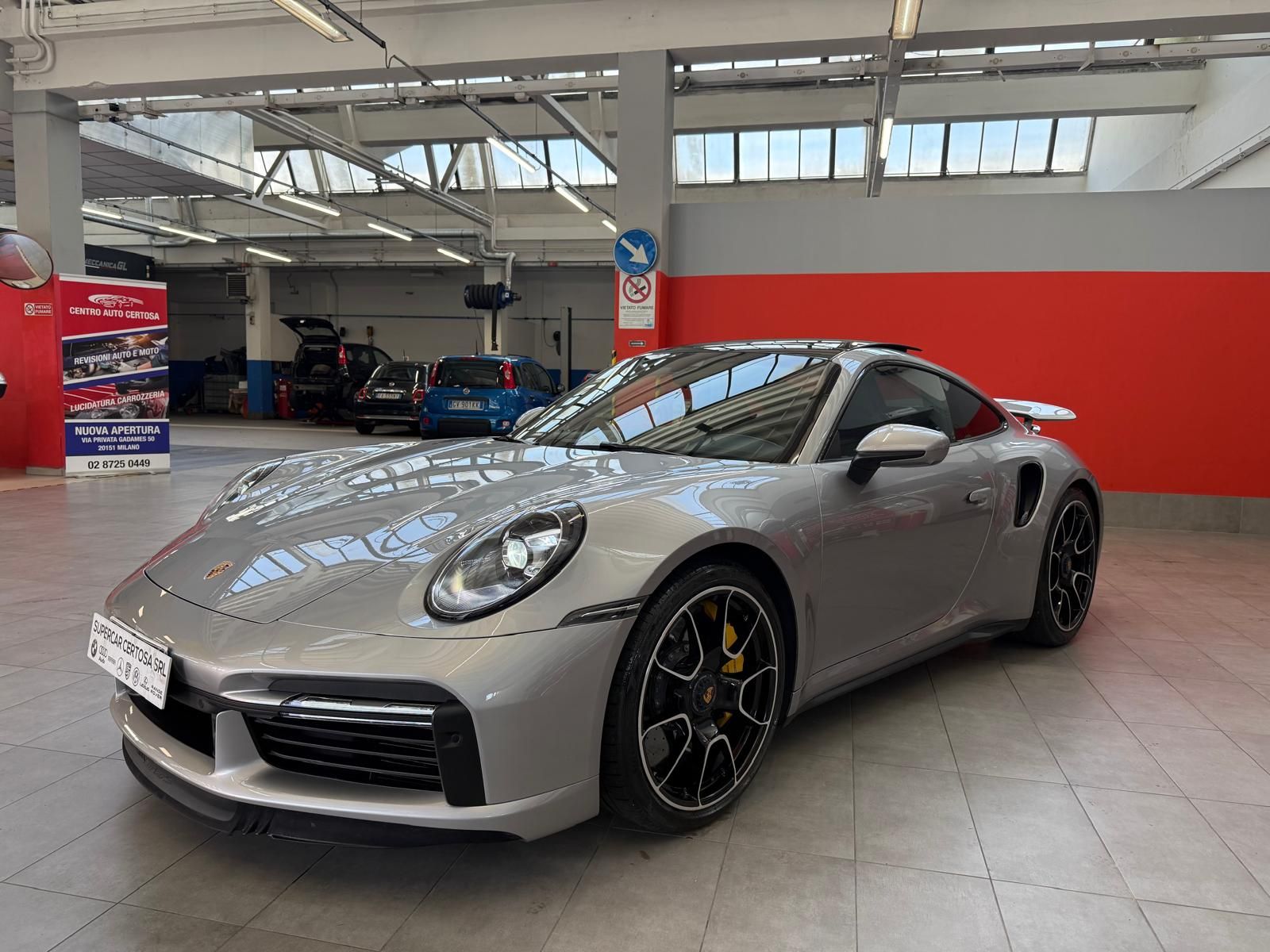 Porsche 911 Turbo S argentata in un'officina con pareti rosse e grigie.