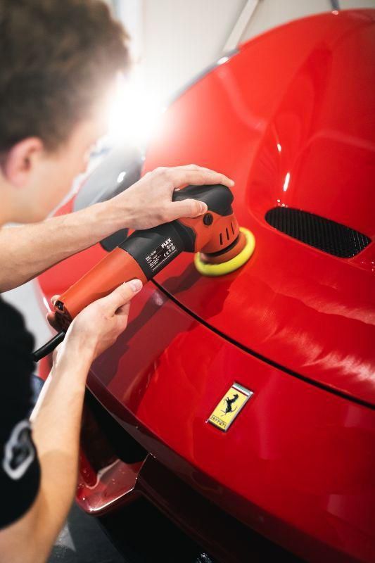 Persona che lucida una Ferrari rosso fuoco con una lucidatrice elettrica in un ambiente ben illuminato.