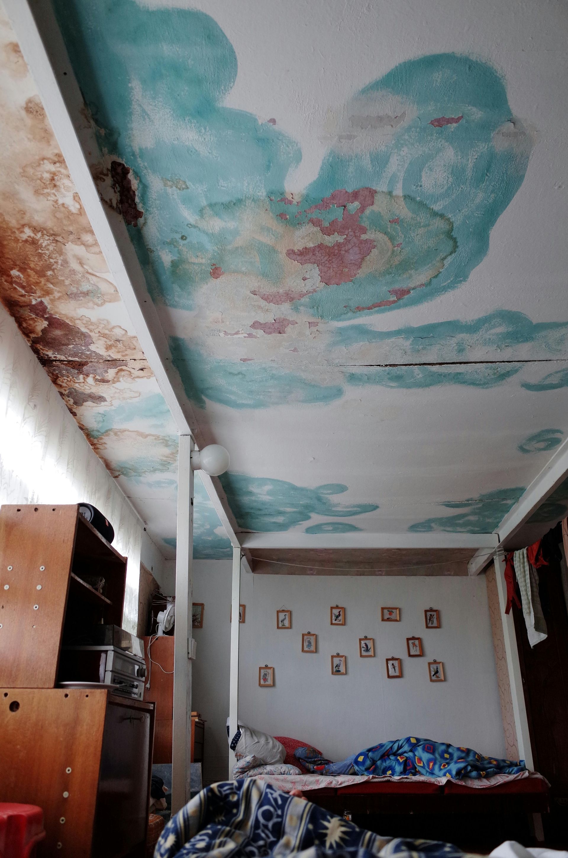 Ceiling with peeling paint resembling clouds in a bedroom; wooden bed frame and wardrobe visible.