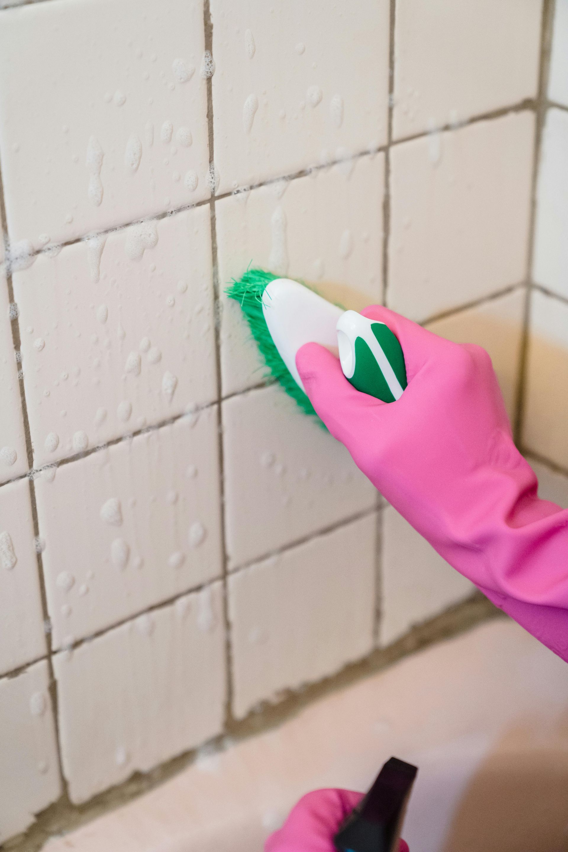 A person wearing pink gloves scrubs a tiled bathroom wall with a green and white brush.