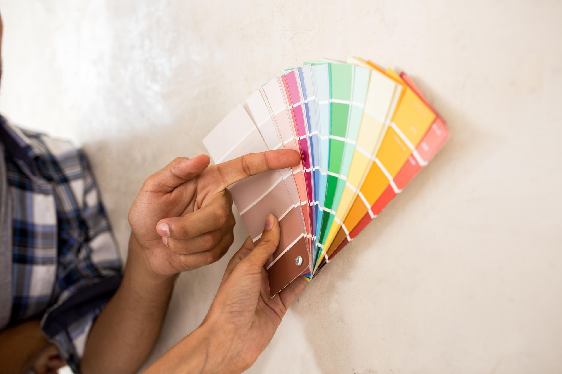Person pointing at pink paint swatch on a color fan against a white wall.