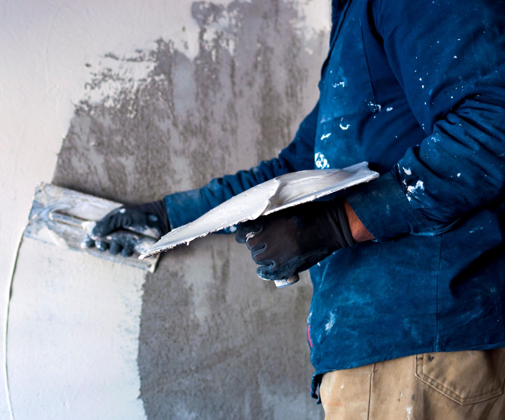 Person in blue jacket applying plaster to a wall with a trowel.