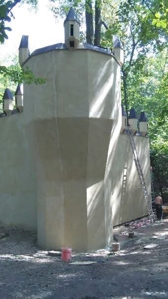 Beige castle-like structure with turrets in a wooded area; ladder leans against it, person nearby.
