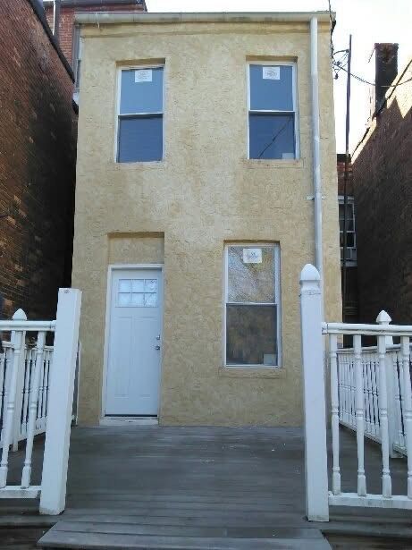 Tan two-story building with a white door and windows.  Wooden deck with railing in front.  Between brick buildings.