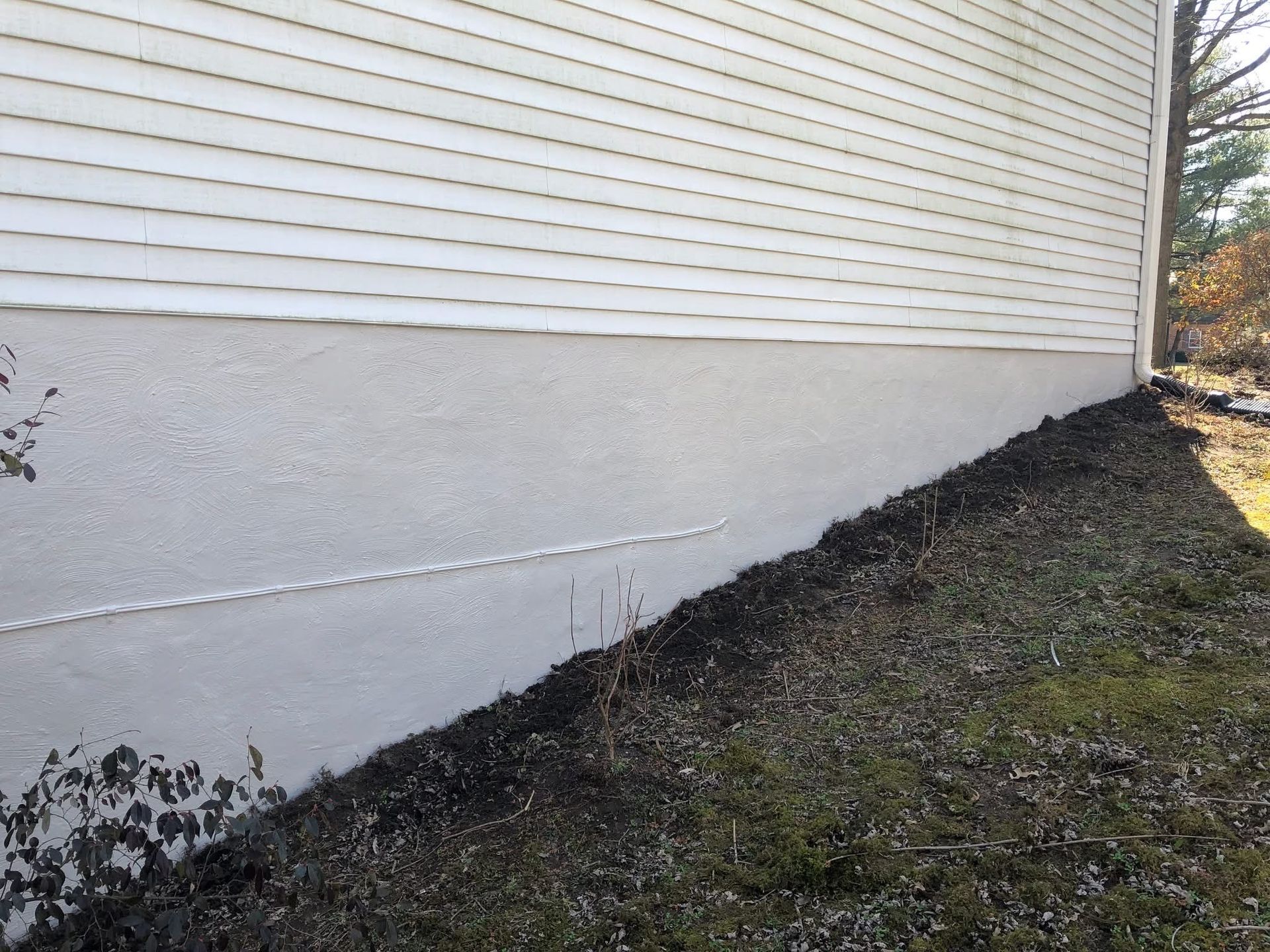 White siding above a textured light-gray foundation. Dark mulch bed with sparse plants sits below.