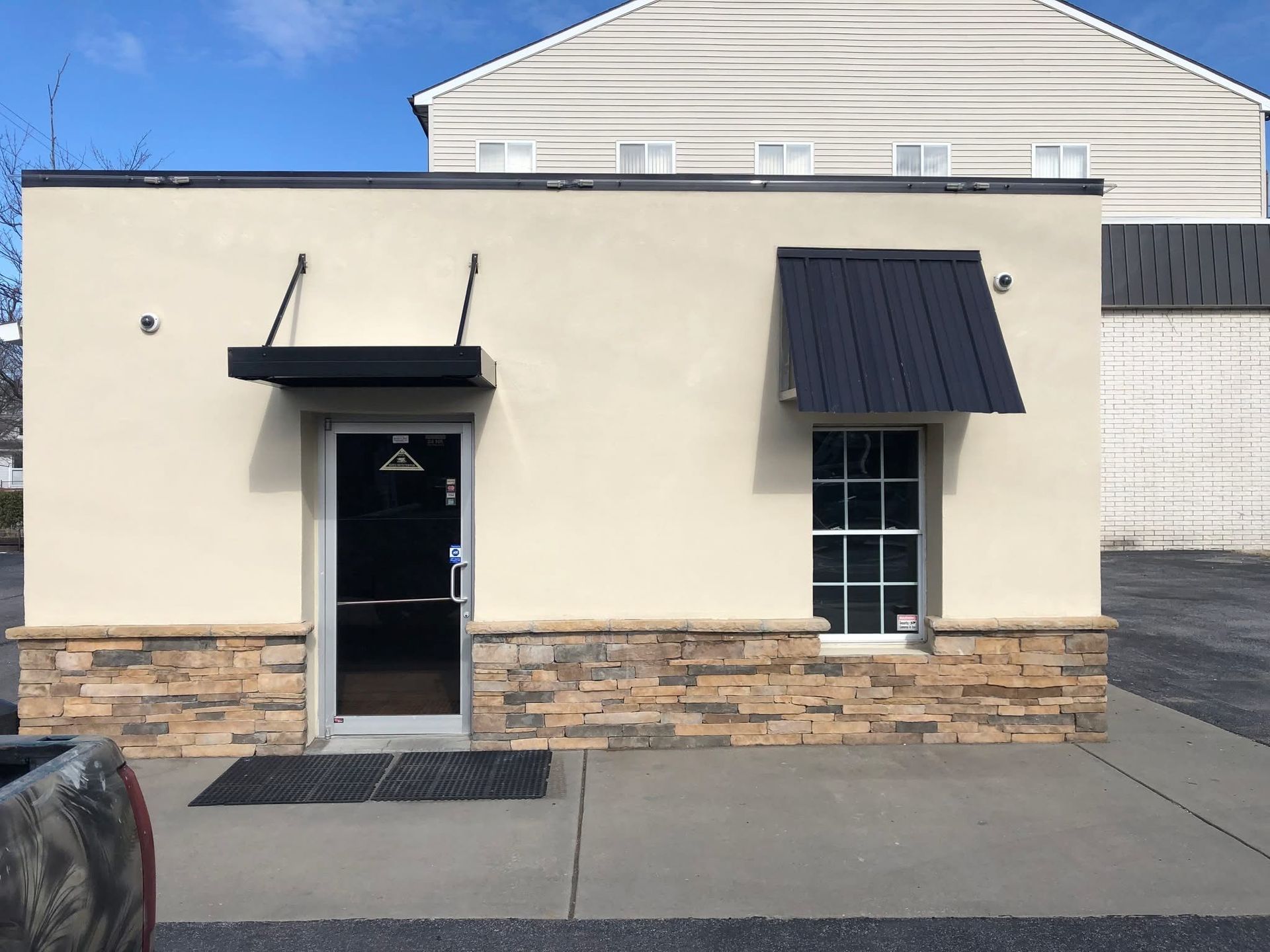 Small, light-beige building with a black door and window. Stone base. Black awnings. Paved ground.