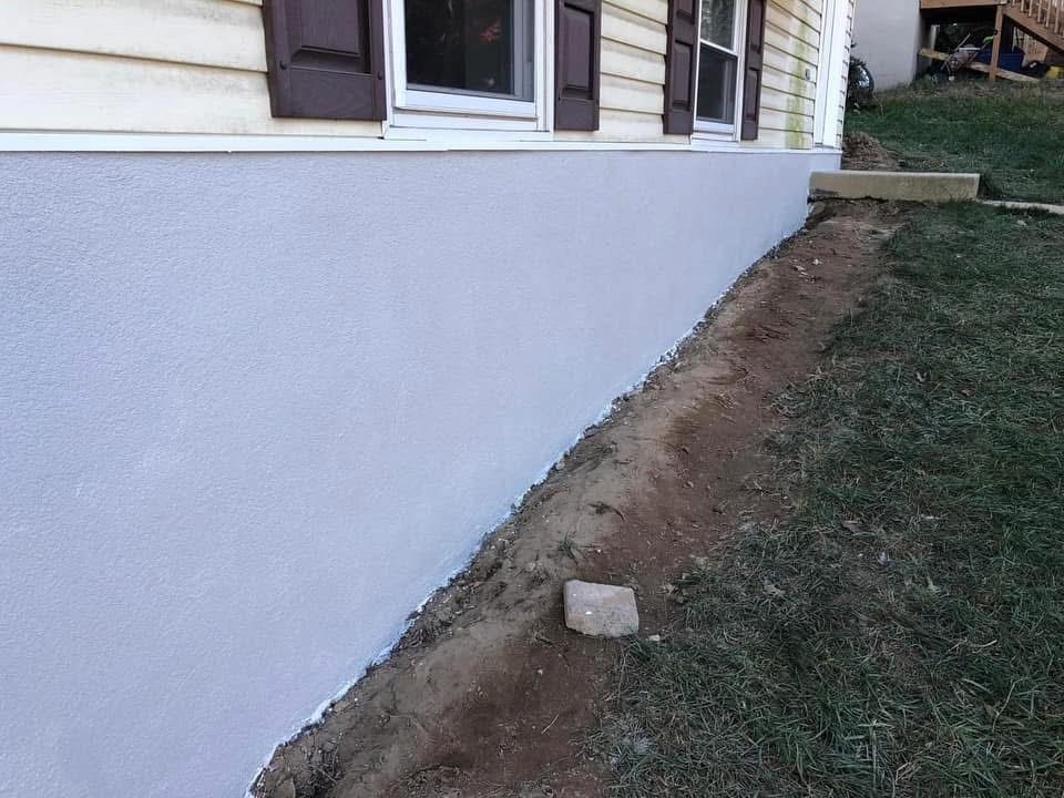 Light purple painted foundation of a house with brown shutters and a dirt patch alongside grass.