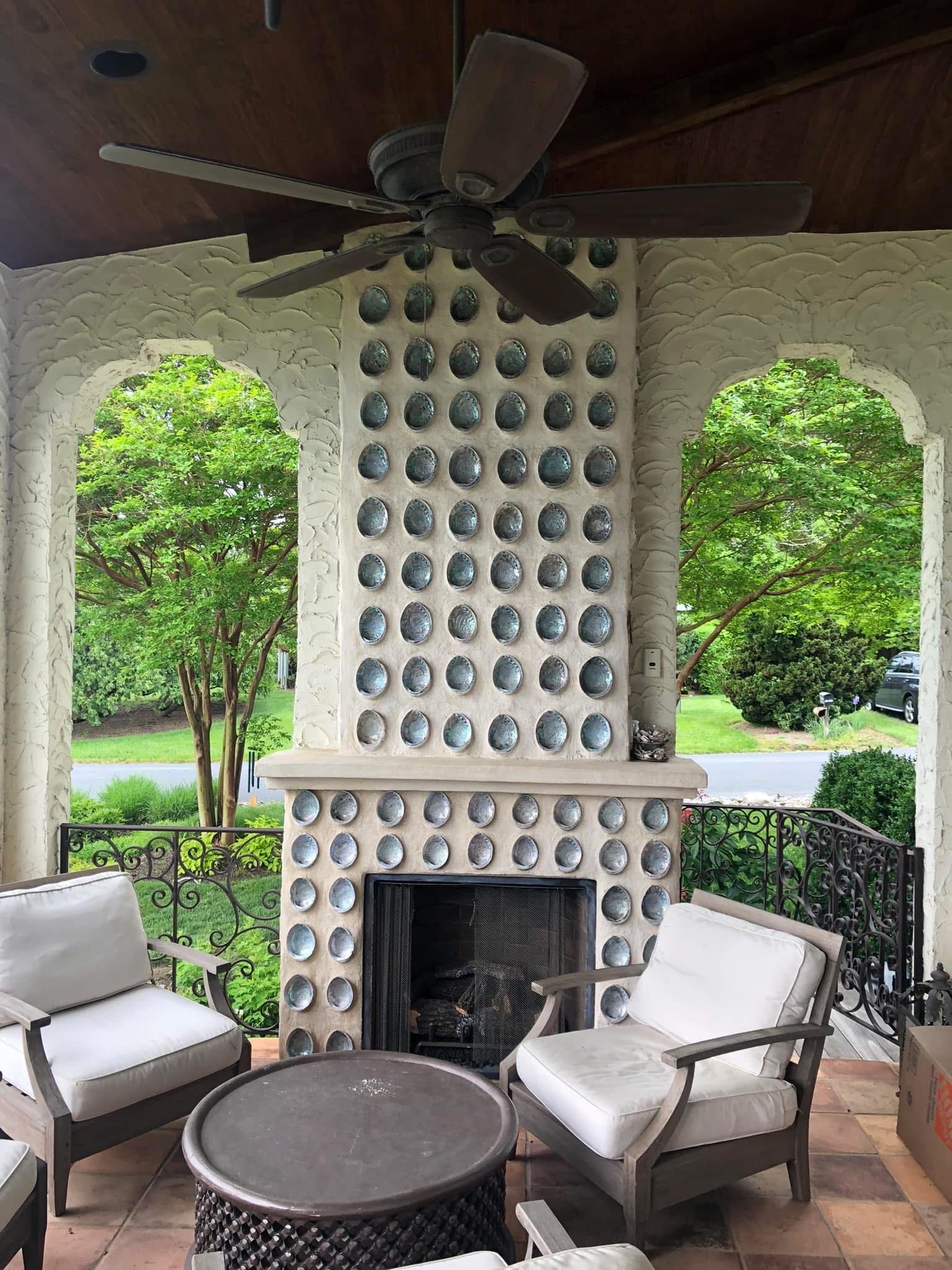 Outdoor patio with fireplace. Arched openings frame trees and street view. Furnished with chairs and a round table.