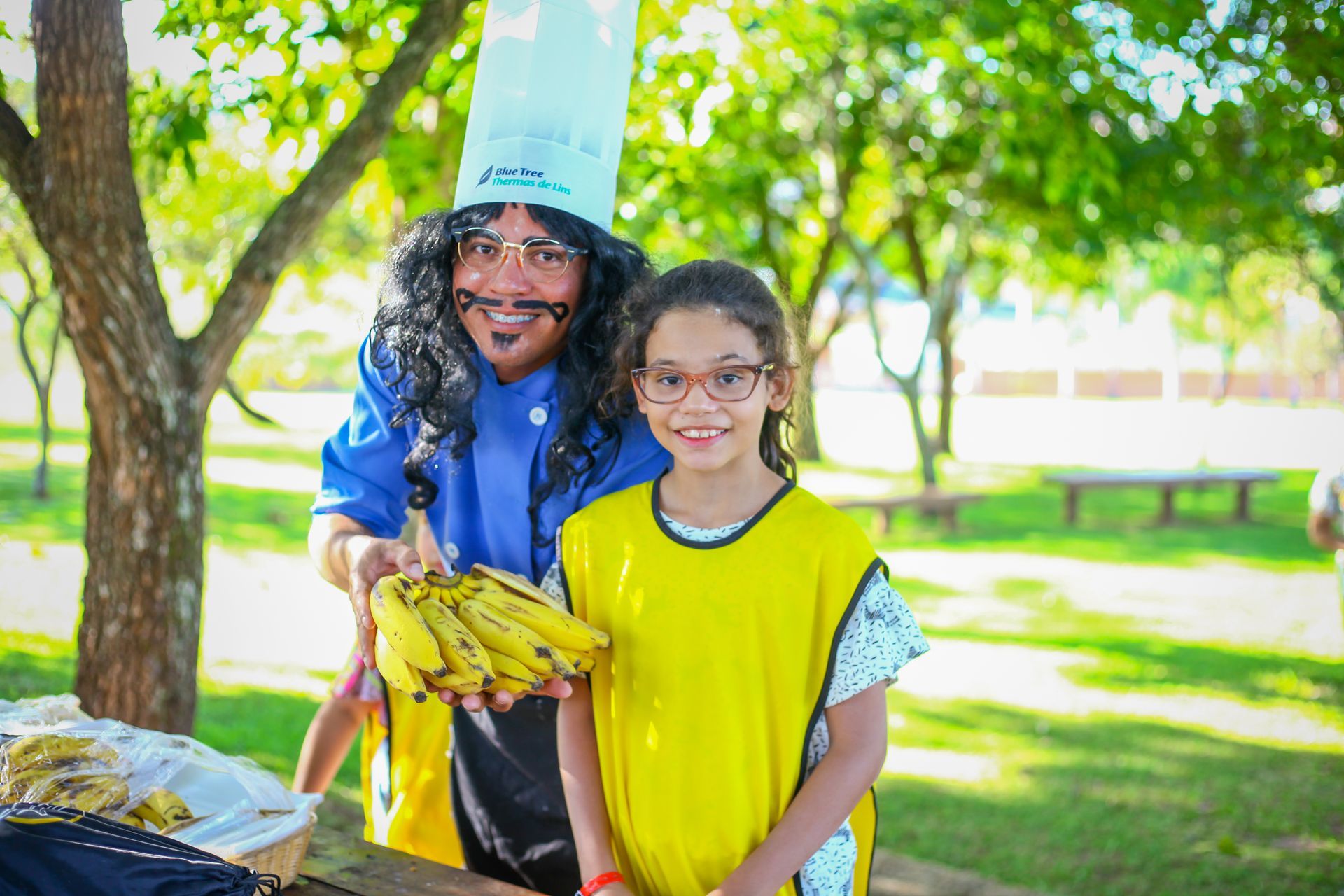 Um homem com chapéu de chef segura um cacho de bananas ao lado de uma garotinha.