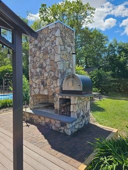 Stone outdoor pizza oven with a chimney, set on a brick patio, near a pool.