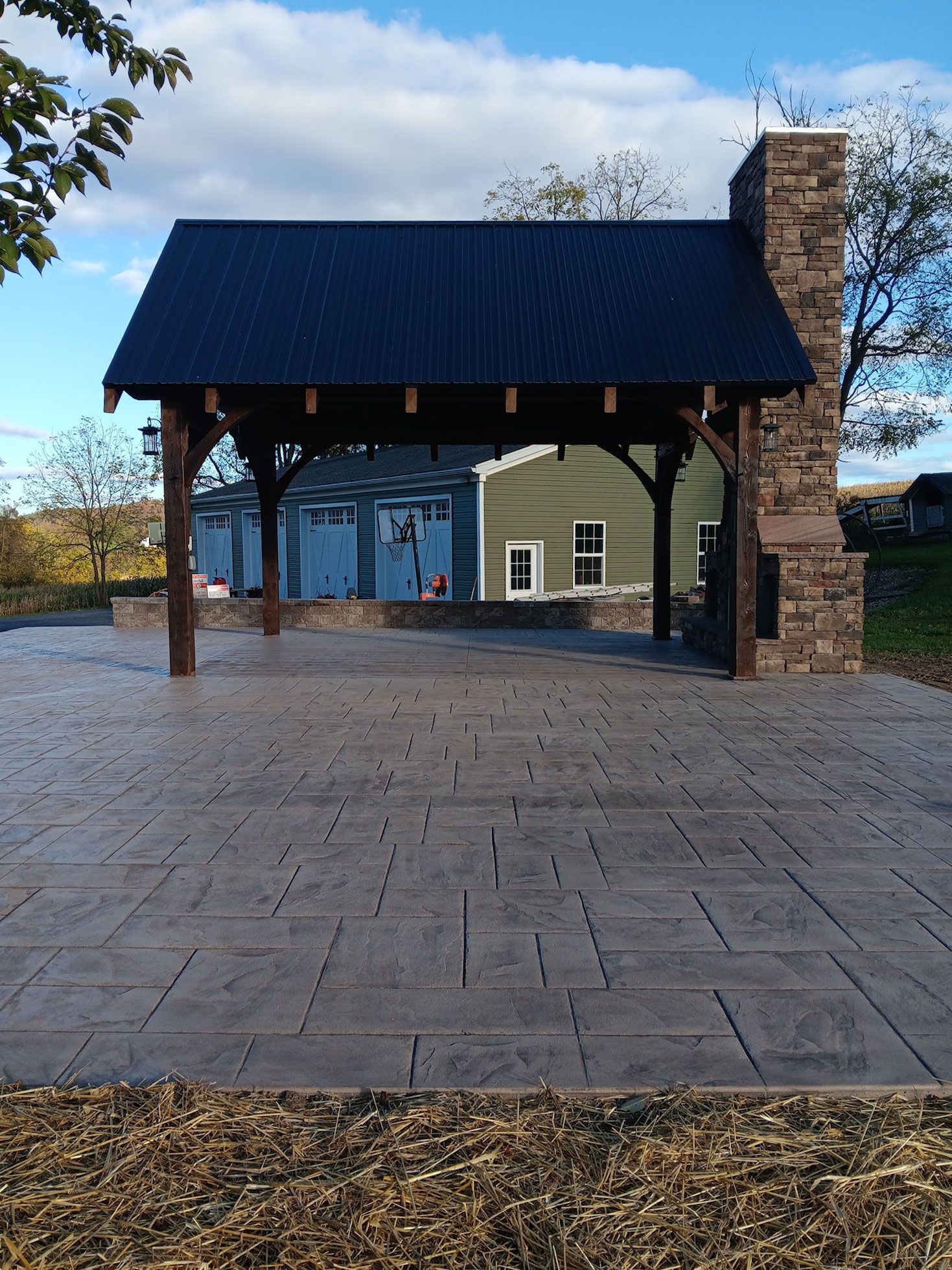 A covered outdoor patio with dark roof, stone chimney, and gray stamped concrete flooring.  Background includes buildings and sky.