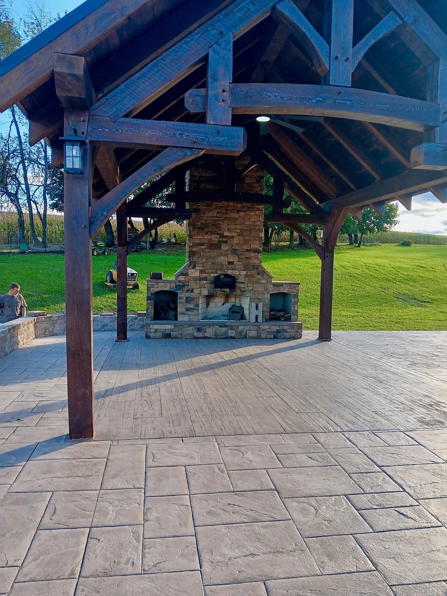 Outdoor brick fireplace under a dark wooden gazebo on a brick patio; green lawn in the background.