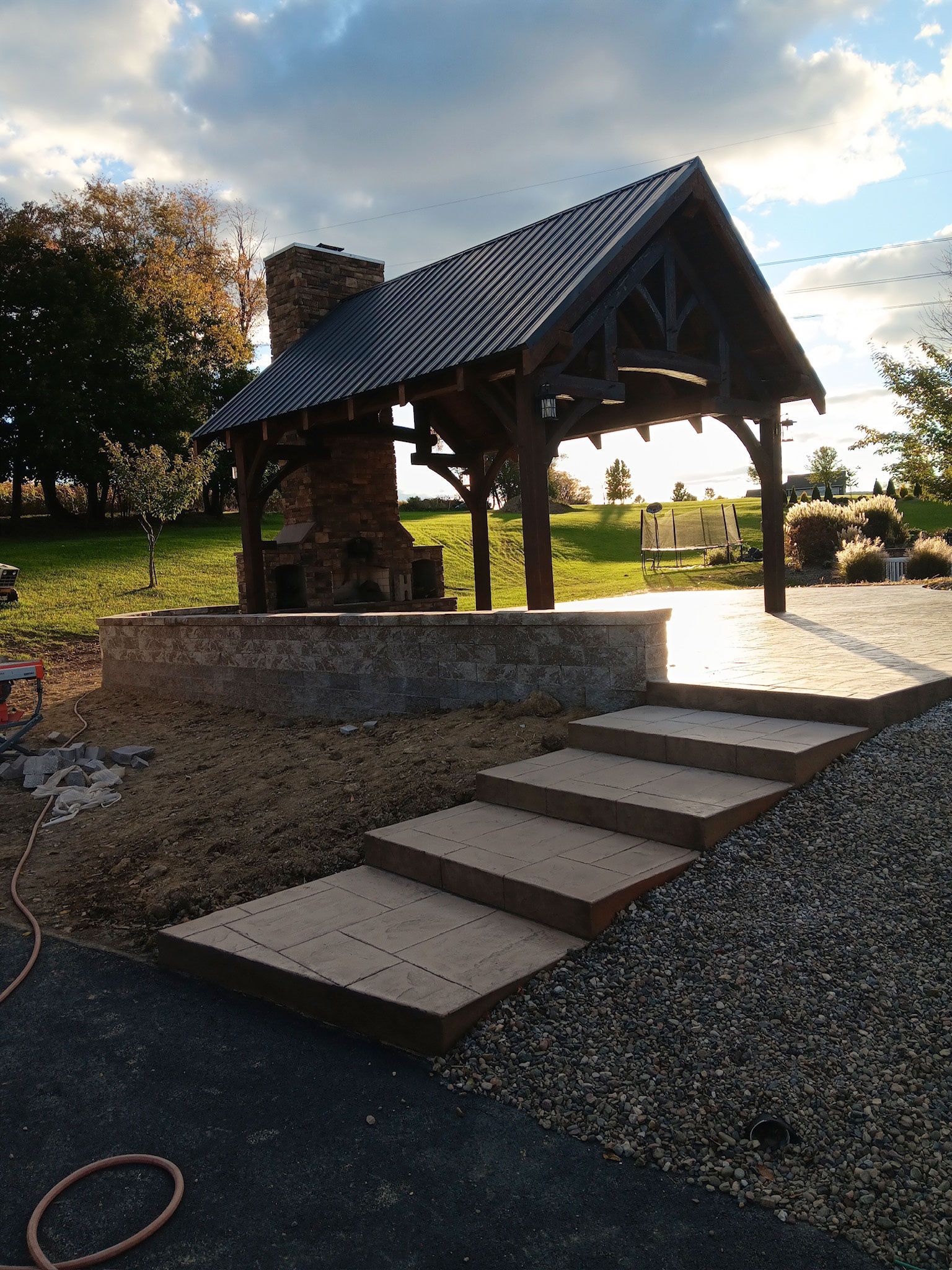 Outdoor stone steps lead to a brick chimney with a wooden roof, set in a grassy yard, lit by sun.