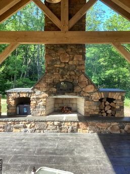 Outdoor stone fireplace with wood storage, set on a patio under a wooden beam structure.