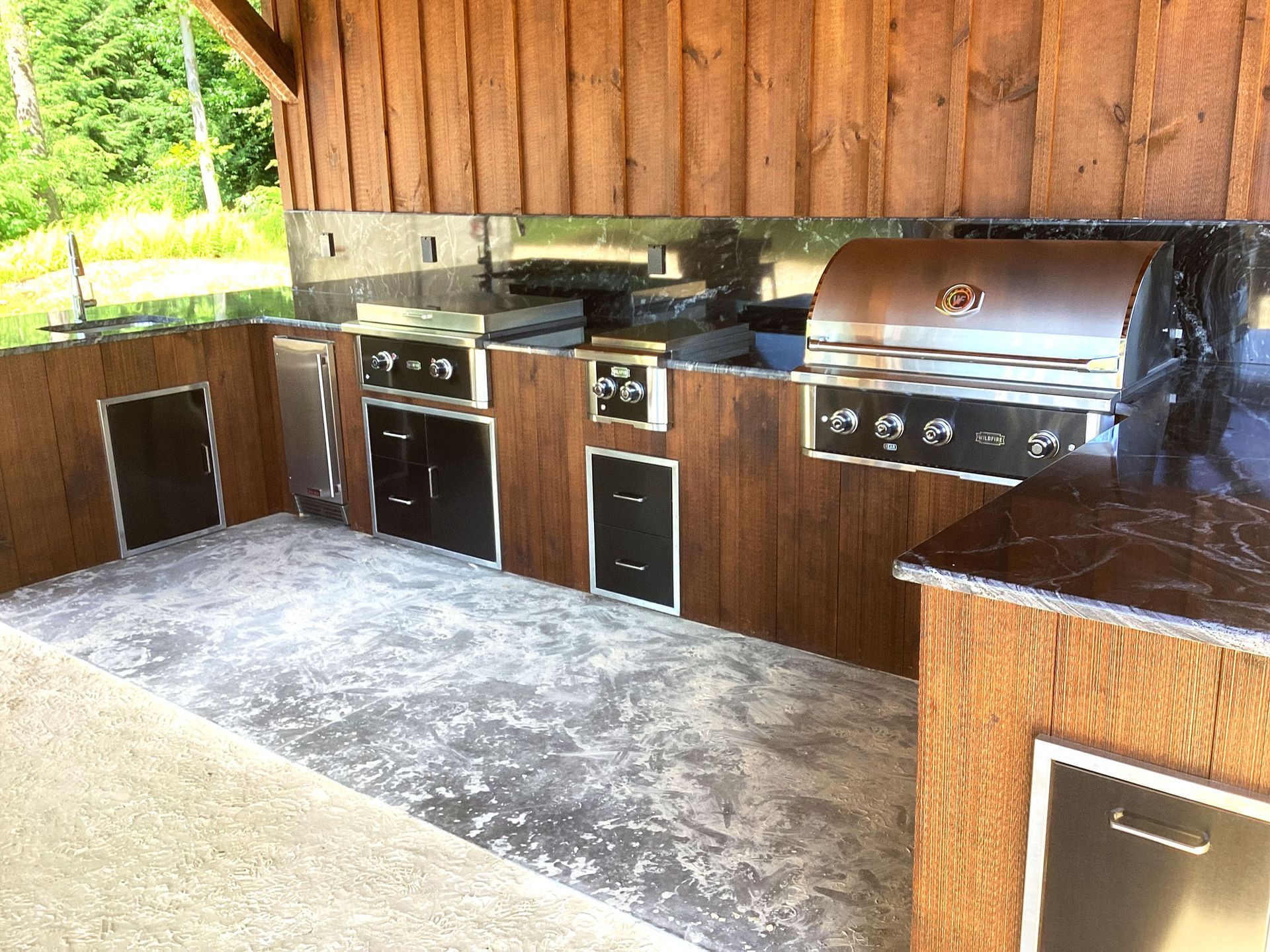 Outdoor kitchen with brown cabinets, concrete floor, and stainless steel grill.