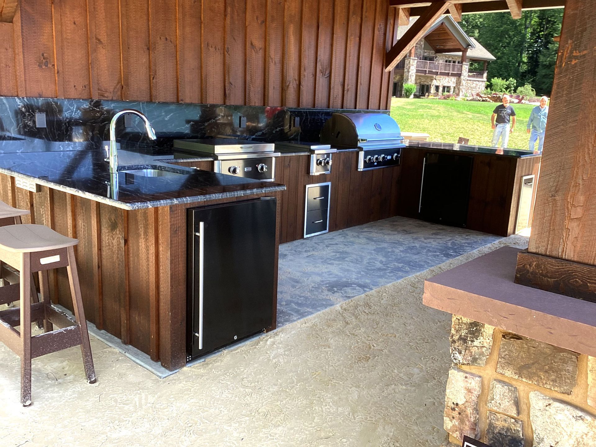 Outdoor kitchen with grill, sink, and fridge on a concrete patio, dark brown wood cabinets, a person in the background.
