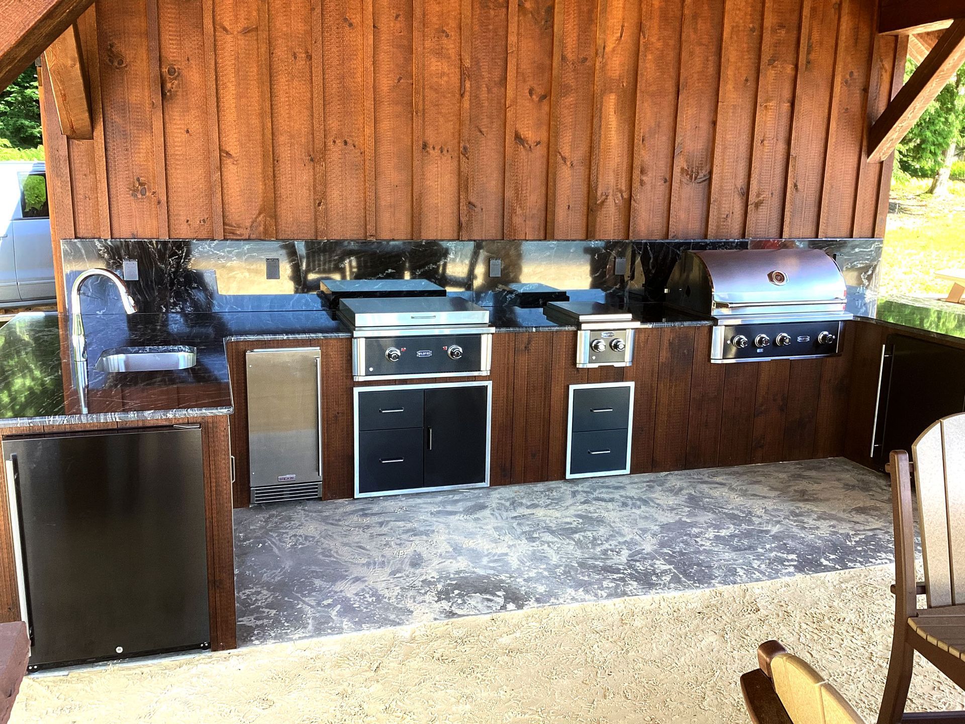 Outdoor kitchen under a wood-paneled structure. Includes a grill, cooktop, storage, and granite countertops.
