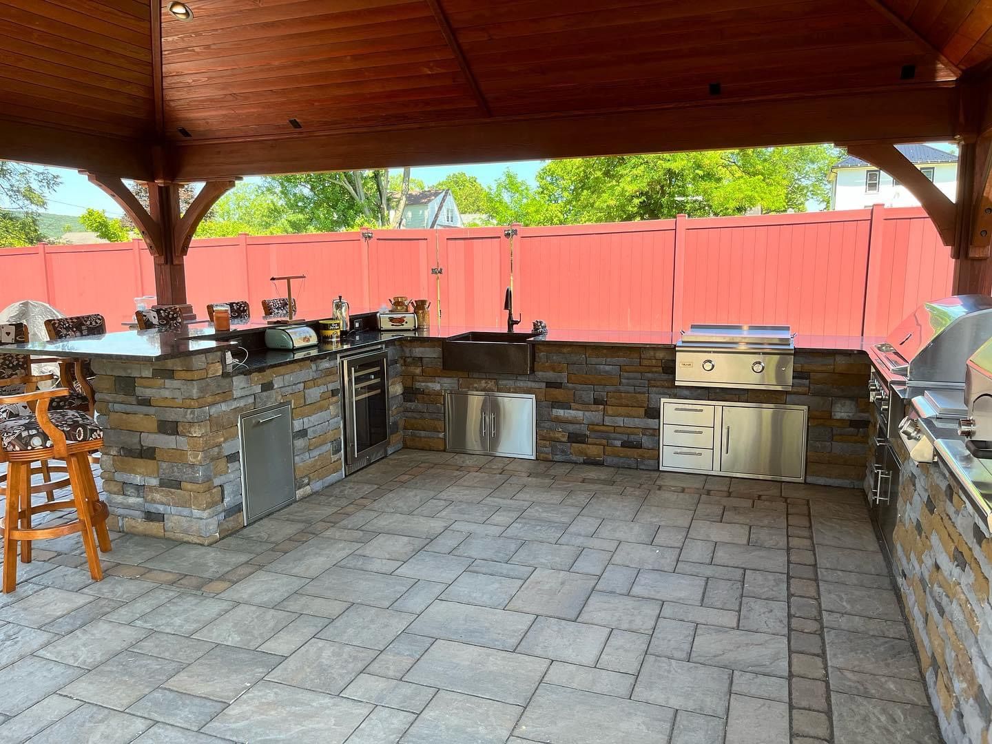 Outdoor kitchen under a wooden gazebo, with stone counters, stainless steel appliances, and a brick patio.