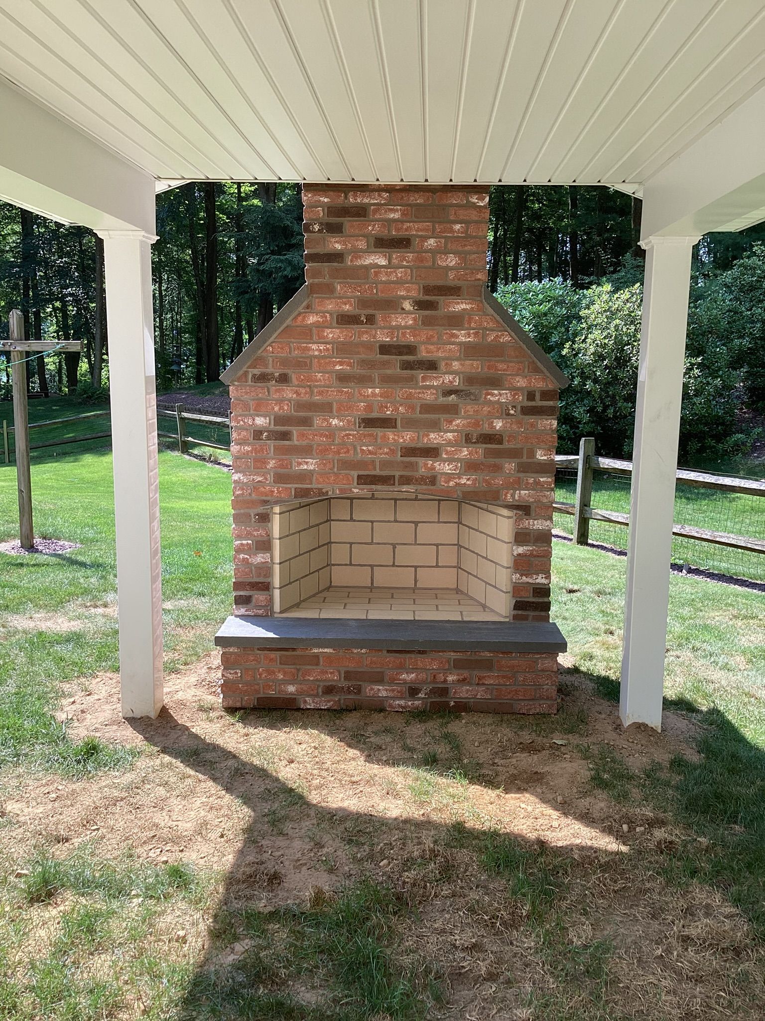 Brick outdoor fireplace under a white-columned covered area with green lawn and trees in the background.