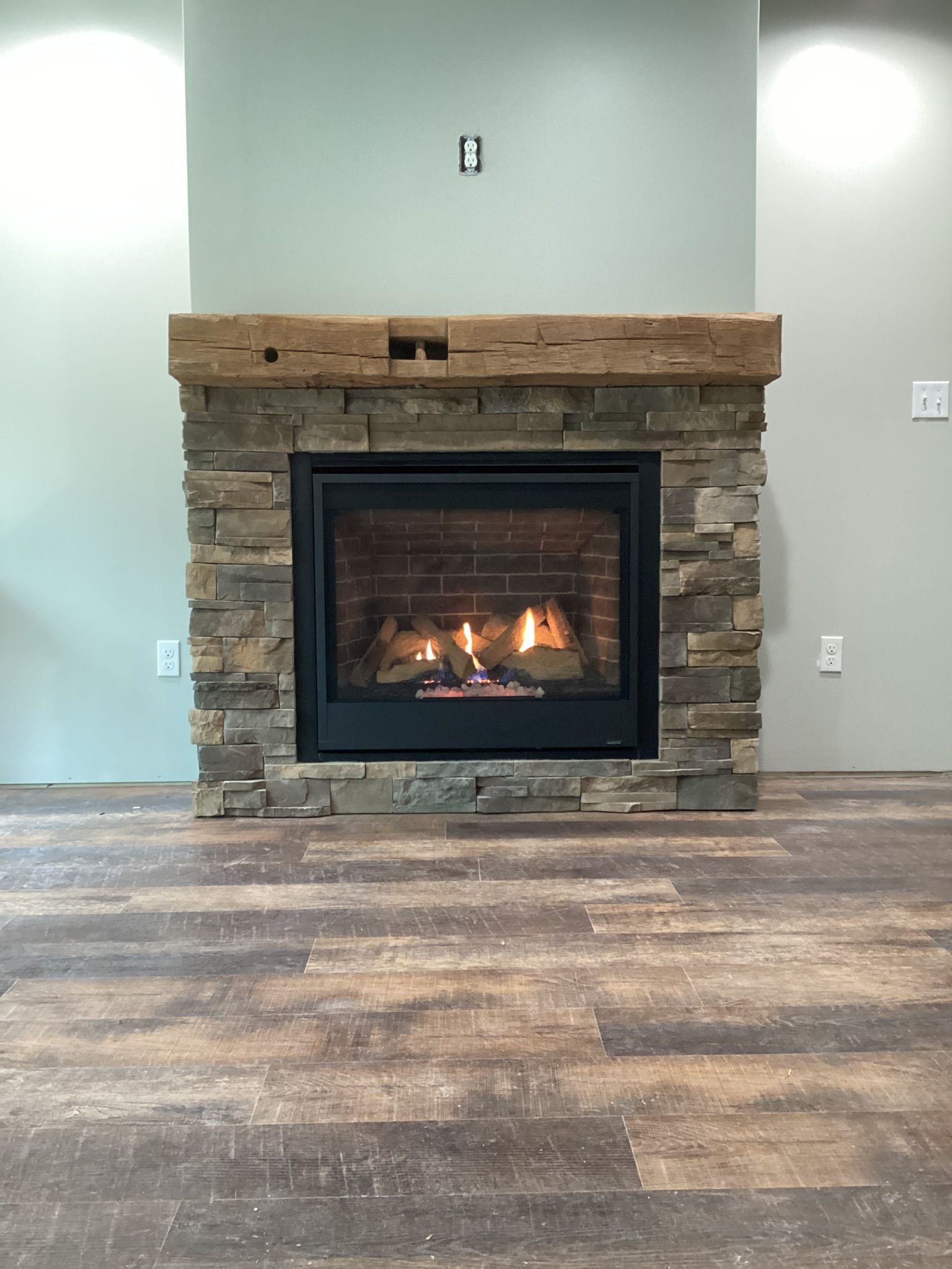 Fireplace with stone surround, wooden mantel, and burning flames. Set in a room with neutral walls and flooring.