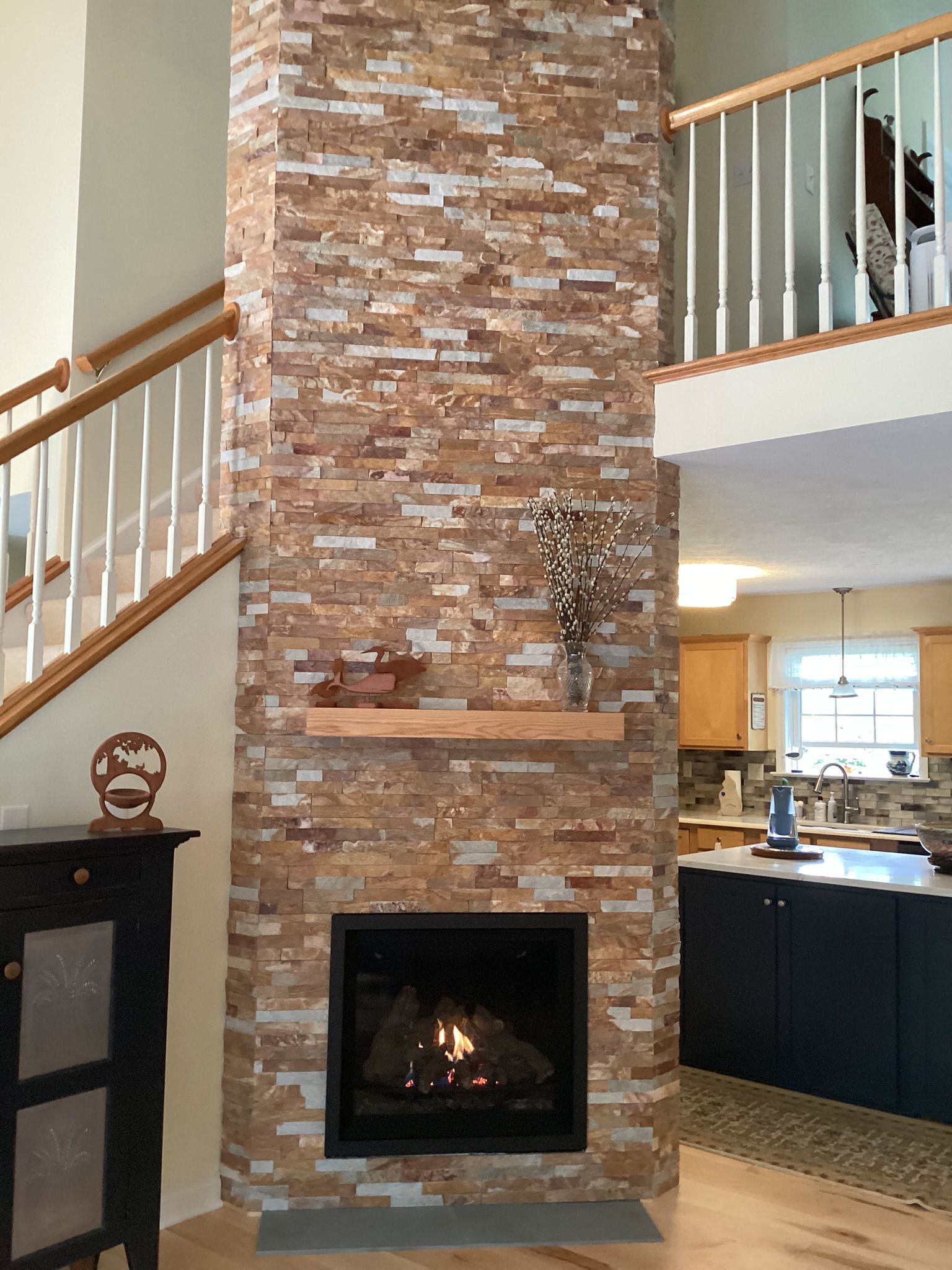 Fireplace with stone facing, wooden mantle, and burning fire. Staircase on the left, kitchen in background.