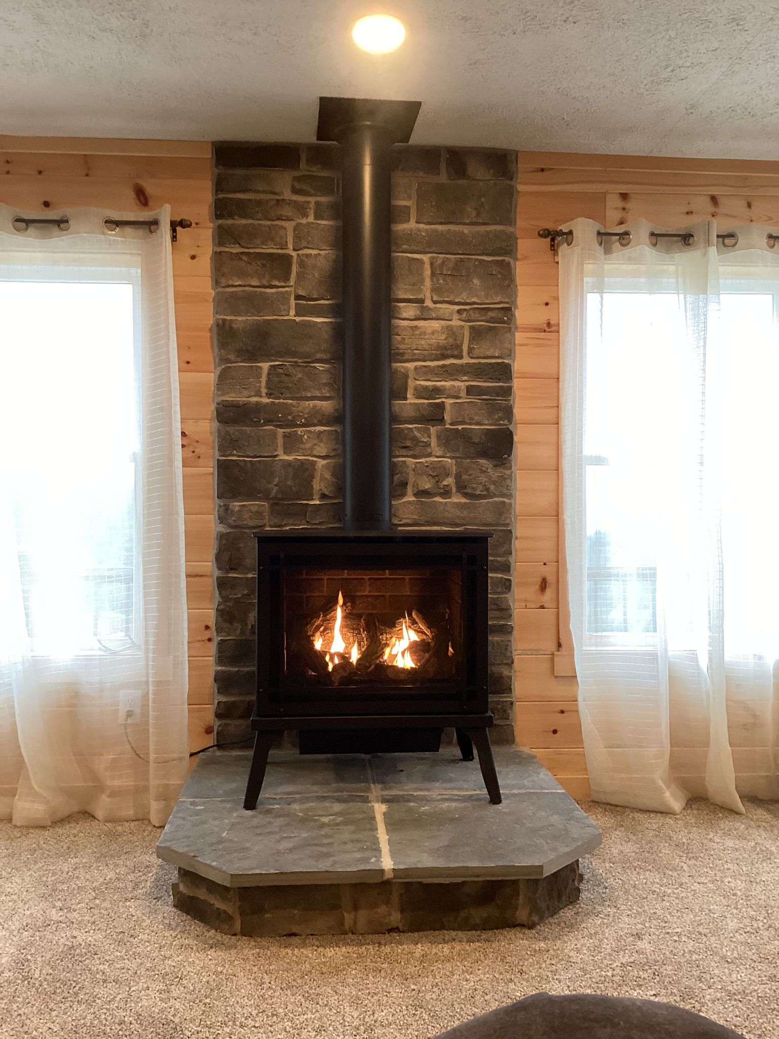 Wood stove burning in a stone hearth, flanked by windows with sheer curtains, in a wood-paneled room.