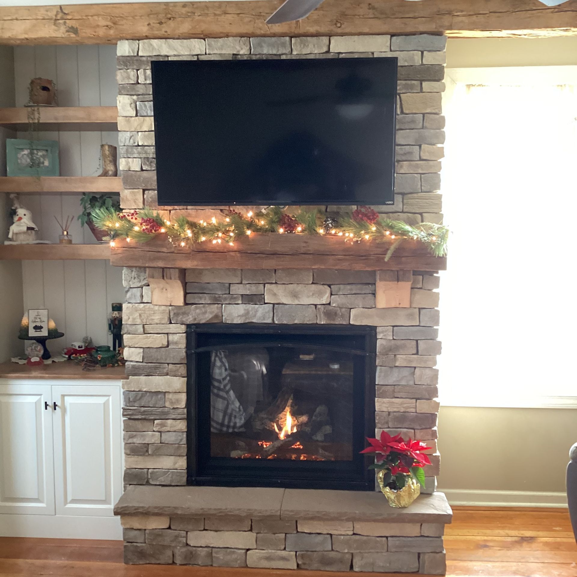 Stone fireplace with lit fire, TV, Christmas garland, and built-in shelves with decor.