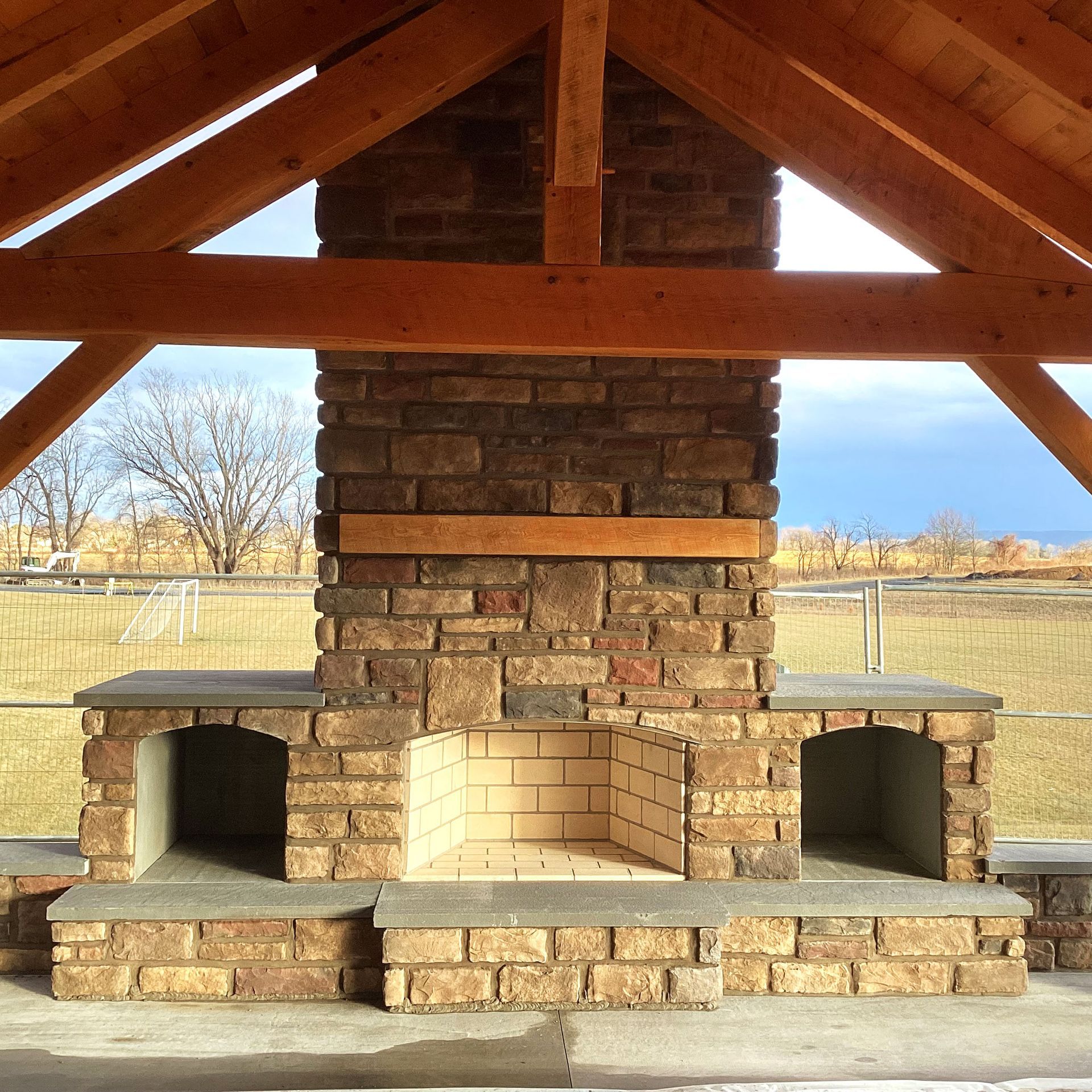 Brick outdoor fireplace under a wooden roof.  Fireplace has two side storage areas.