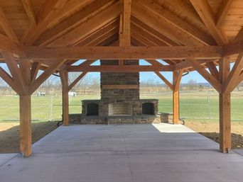 Wooden pavilion with stone fireplace on a concrete patio, overlooking a grassy field.