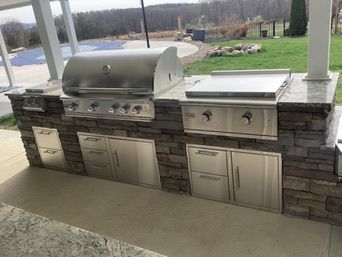 Outdoor kitchen with stainless steel grill and griddle, stone facade, and cabinets.