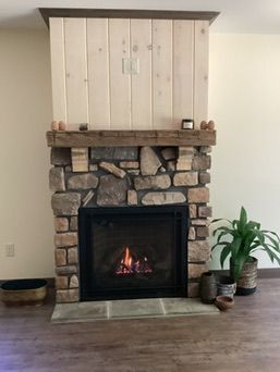 Fireplace with stone facade, wooden mantel, and a lit fire. There are potted plants nearby.