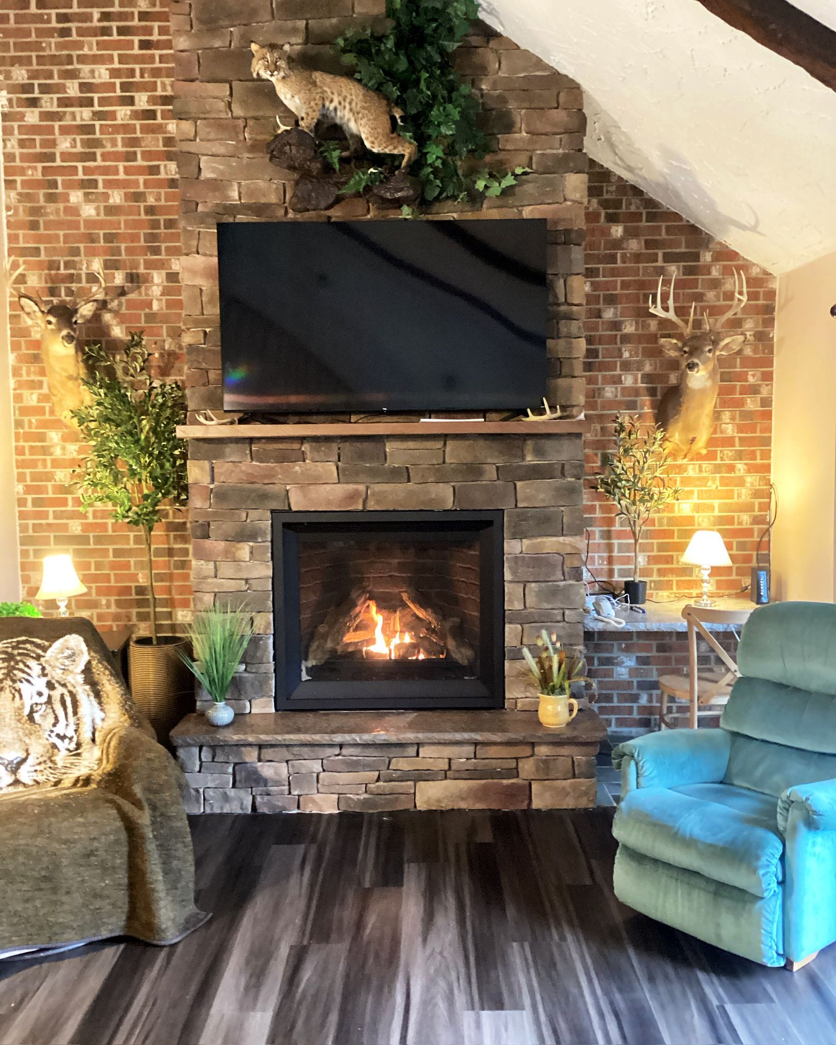 Cozy living room with brick fireplace, TV, taxidermy, and green armchairs.