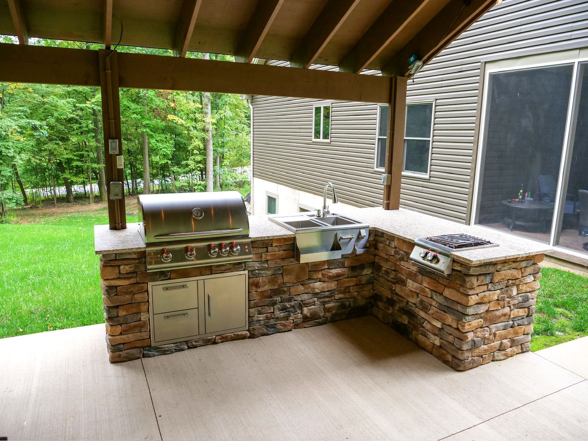 Outdoor kitchen with stone facade, grill, sink, cooktop under a wooden pergola.